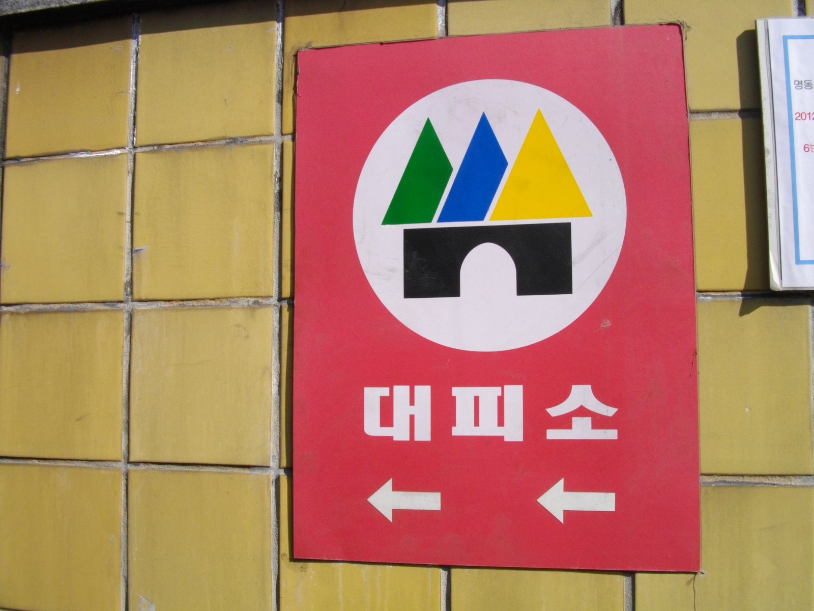 노란 타일 벽에 붙은 빨간 민방위 대피소 유도표지판. 흰 원 안에 녹색·파란색·노란색 삼각형과 검은 터널 모양이 있고, 아래에 흰 글씨로 대피소와 왼쪽 화살표 두 개가 있다