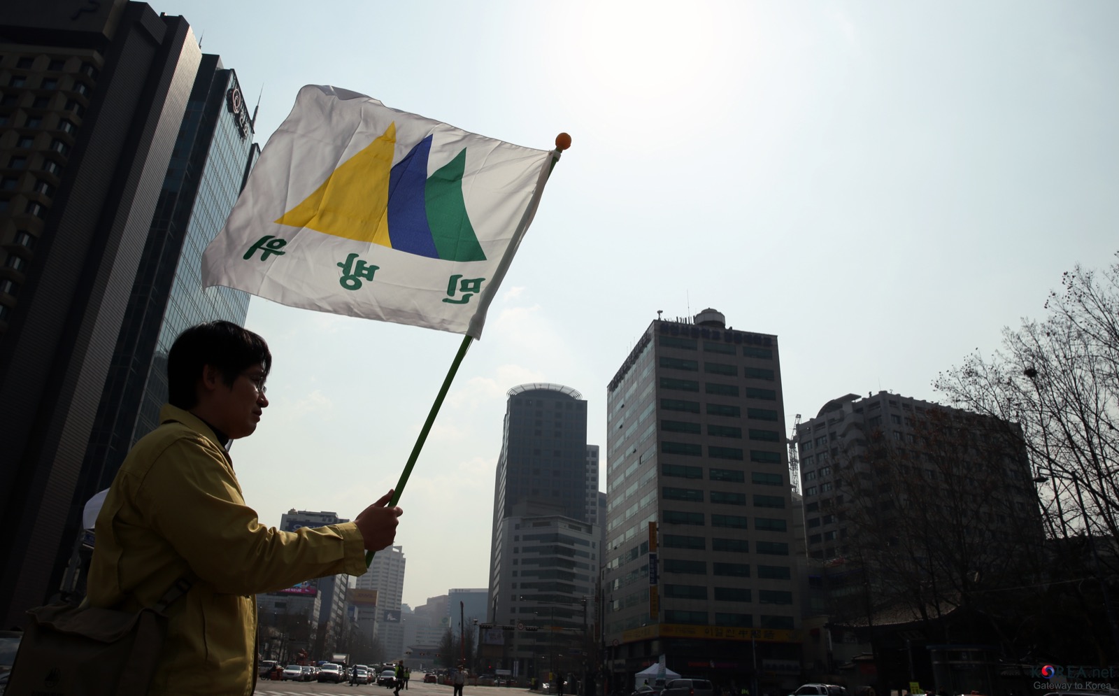서울 도심 교차로에서 노란 민방위복을 입은 사람이 민방위 로고가 그려진 깃발을 들고 서 있고 뒤로 고층 건물이 보인다